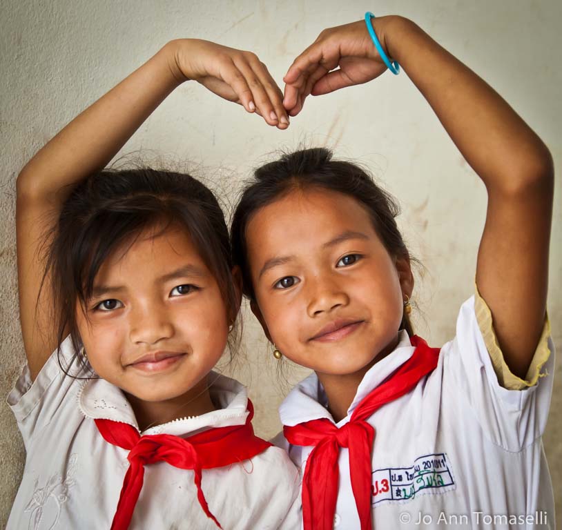 A portrait  photograph by Award Winning Photographer Jo Ann Tomaselli of two schoolgirls in Laos wearing white shirts and red scarves show that they're best friend by linking their hands and forming a heart around themselves. Buy Best Fine Art Photography at  http://jo-ann-tomaselli.artistwebsites.com/